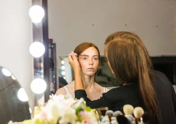 Prozess der Herstellung von Make-up. Make-up-Artist arbeitet mit Pinsel auf Modell Gesicht. Porträt einer jungen Frau mit rotem Haar im Interieur eines Schönheitssalons. Ton auf die Haut auftragen. — Stockfoto