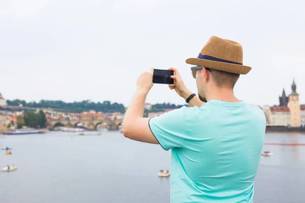 Bir genç adam turist nehir ve şehir önünde smartphone ile fotoğraf çekme arkadan görünüm — Stok fotoğraf