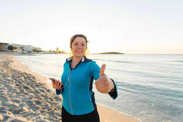 Concept van sport, fitness, gezonde levensstijl en running - gemotiveerd sportieve vrouw doen duimschroef opwaarts succes gebaar na buiten training op het strand — Stockfoto