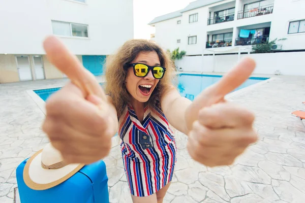 Gelukkig meisje duimen opdagen op zomervakantie op de achtergrond van zwembad, gekleed gestreepte jurk en zonnebril. — Stockfoto