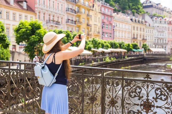 Mujer tomando fotos con smartphone. Elegante viajero de verano mujer en sombrero con cámara al aire libre en la ciudad europea, casco antiguo de Karlovy Vary en el fondo, República Checa, Europa — Foto de Stock