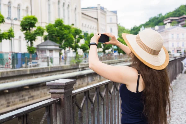 Mujer tomando fotos con smartphone. Elegante viajero de verano mujer en sombrero con cámara al aire libre en la ciudad europea, casco antiguo de Karlovy Vary en el fondo, República Checa, Europa — Foto de Stock