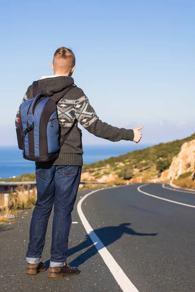 Joven caucásico visto por detrás llevando una mochila haciendo autostop en un camino menor, con el pulgar hacia arriba — Foto de Stock