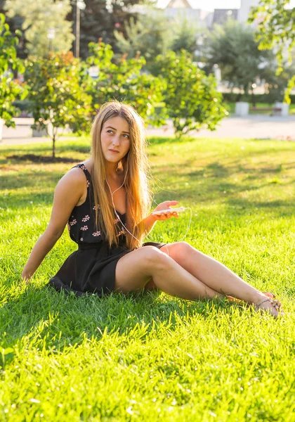 Hermosa chica sentada en la hierba en el parque y escuchar música —  Fotos de Stock