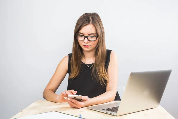 Νεαρή γυναίκα κάθεται στο γραφείο στο τραπέζι και χρησιμοποιώντας το smartphone. Στο γραφείο είναι laptop. Γυναίκα ανάλυση δεδομένων. Φοιτητής Μαθαίνοντας διαδικτυακά. — Φωτογραφία Αρχείου