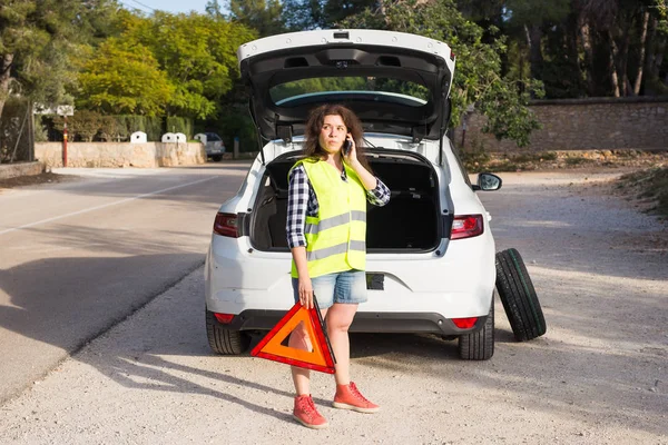 Broken down car with warning triangle. Woman standing alongside her broken down car on the road and making call while waiting for emergancy assistance