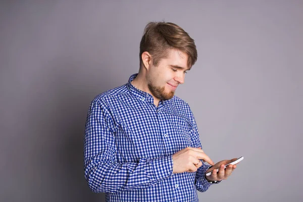 Молодой человек пишет смс на смартфоне, изолированном на сером фоне. Улыбающийся парень держит смартфон и смотрит на него . — стоковое фото