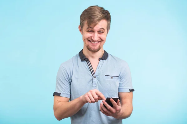 Hombre divertido mensaje de texto en el teléfono inteligente aislado en fondo azul. Sonriente tipo sosteniendo el teléfono inteligente y mirándolo . — Foto de Stock