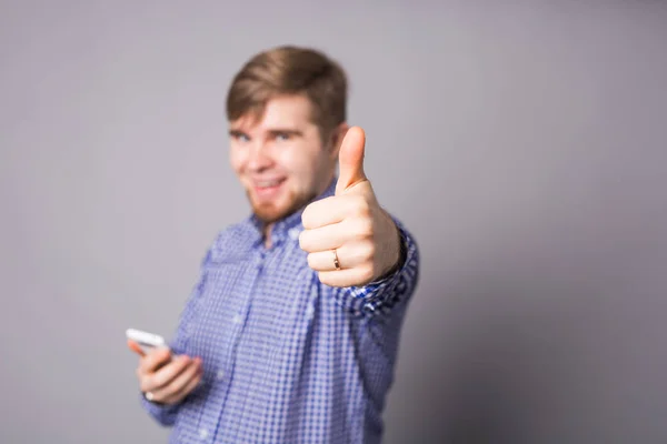 Junger gutaussehender Mann hält sein Smartphone und zeigt gestenreich den Daumen nach oben — Stockfoto