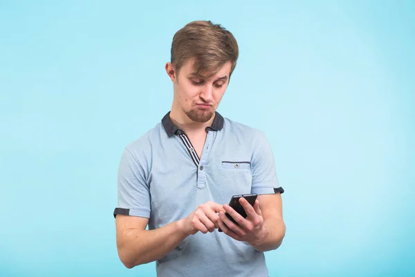 Забавный человек пишет смс на смартфоне, изолированном на синем фоне. Улыбающийся парень держит смартфон и смотрит на него . — стоковое фото