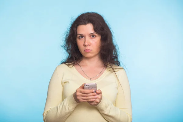 在蓝色背景下手持智能手机看摄像头的不满的不开心的女人 — 图库照片