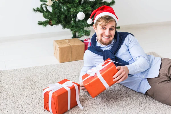 Kerstmis en feestdagen concept - Gelukkig man in de kerstman hoed openen van een geschenk thuis in de woonkamer — Stockfoto