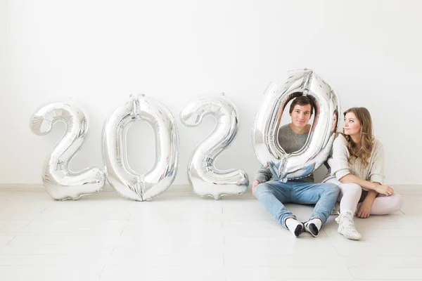 Vacaciones, concepto festivo y de fiesta - Pareja divertida sentada en un piso cerca de globos plateados 2020 sobre fondo blanco. Celebración de Año Nuevo — Foto de Stock