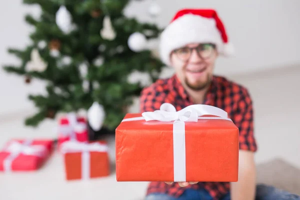 Concepto de Navidad y vacaciones - Hombre divertido en sombrero de santa celebración de un regalo en casa en la sala de estar —  Fotos de Stock