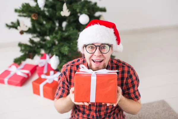 Concepto de Navidad y vacaciones - Hombre divertido en sombrero de santa celebración de un regalo en casa en la sala de estar — Foto de Stock