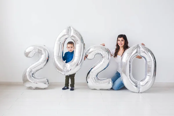 Año nuevo, celebración y concepto de vacaciones - madre e hijo sentados cerca de signo 2020 de globos de plata para el año nuevo en el fondo de la habitación blanca —  Fotos de Stock