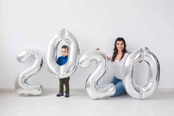 Año nuevo, celebración y concepto de vacaciones - madre e hijo sentados cerca de signo 2020 de globos de plata para el año nuevo en el fondo de la habitación blanca — Foto de Stock