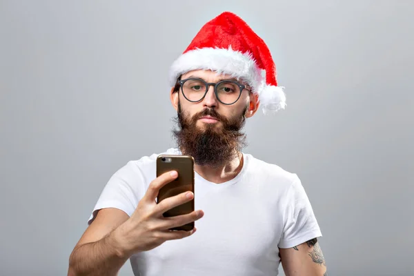 Navidad, vacaciones, tecnología y concepto de la gente - hombre barbudo guapo en sombrero de santa tomar foto selfie con teléfono inteligente sobre fondo gris —  Fotos de Stock
