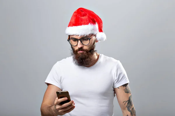 Navidad, vacaciones, tecnología y concepto de la gente - hombre barbudo guapo en sombrero de santa tomar foto selfie con teléfono inteligente sobre fondo gris — Foto de Stock