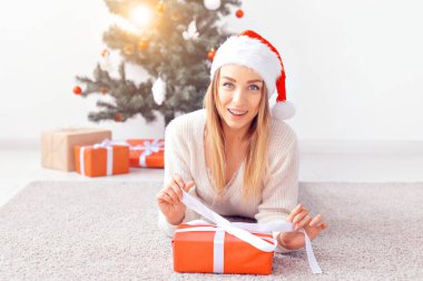 Güzel sarışın kadın sıcak örgü bir kazak giyiyor, oturma odasındaki dekore edilmiş Noel ağacının yakınındaki bir halıya uzanıyor. Ağacın altında bir sürü hediye.