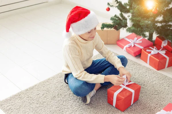 休日、クリスマス、子供時代と人々のコンセプト-サンタの帽子の幸せなティーンの男の子がクリスマスツリーの背景にギフトボックスを開きます — ストック写真