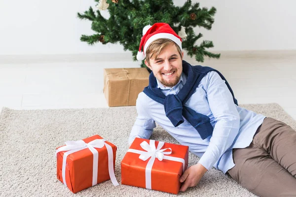 Concepto de Navidad y vacaciones - Hombre feliz en sombrero de santa apertura de un regalo en casa en la sala de estar —  Fotos de Stock