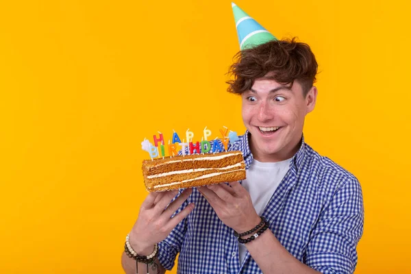 Pozitív vicces fiatal fickó egy sapkát és egy házi készítésű sütemény a kezében pózol egy sárga háttér. Évforduló-és születésnapi koncepció. — Stock Fotó