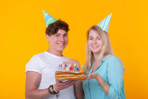 Young cheerful students charming girl and nice guy in greeting paper caps holding a cake. Concept of congratulations on the birthday and anniversary