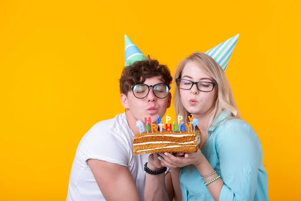 Забавная молодая пара в бумажных кепках и с тортом делают глупое лицо и желают счастливого дня рождения, стоя на желтом фоне. Концепция поздравлений и розыгрышей . — стоковое фото