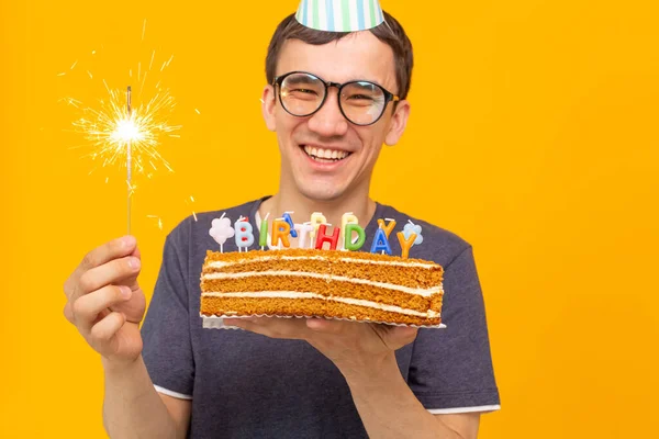 Positivo divertente ragazzo asiatico con un tappo e una candela accesa e una torta fatta in casa tra le mani in posa su uno sfondo giallo. Anniversario e concetto di compleanno . — Foto Stock