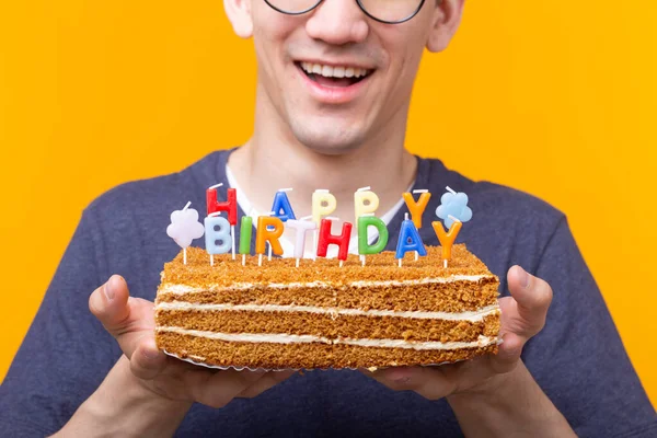 Позитивний смішний молодий азіатський хлопець з шапочкою і палаючою свічкою і домашній торт в руках позує на жовтому тлі. Концепція ювілею та дня народження . — стокове фото