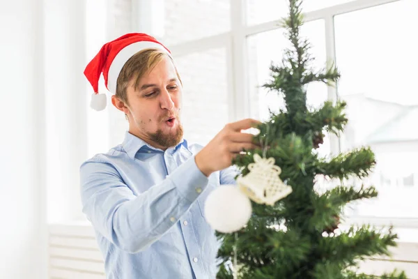Szczęśliwy człowiek ozdabiający choinkę w domu kapeluszem Mikołaja. Człowiek dekorowanie drzewa z bombkami podczas ferii zimowych. — Zdjęcie stockowe
