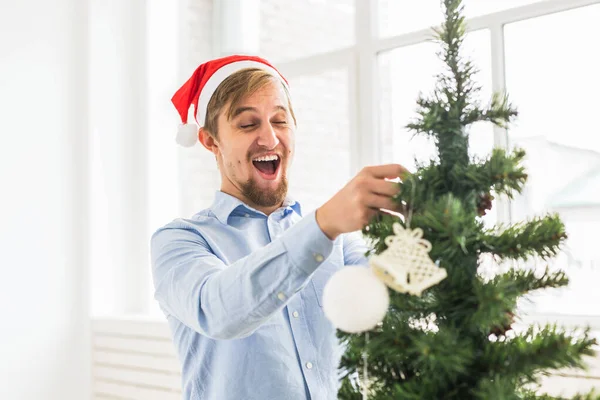 Šťastný muž zdobí vánoční stromek doma kloboukem Santa Clause. Muž zdobení strom s ozdoby během zimní dovolené. — Stock fotografie