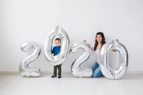 Año nuevo, celebración y concepto de vacaciones - madre e hijo sentados cerca de signo 2020 de globos de plata para el año nuevo en el fondo de la habitación blanca — Foto de Stock