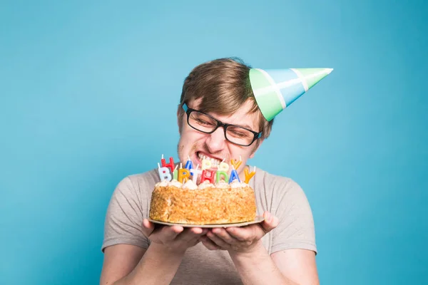 Divertido macho en papel sombrero de felicitación tratando de morder un pastel con un feliz cumpleaños velas de pie sobre un fondo azul . —  Fotos de Stock