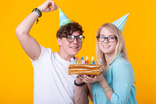 종이 모자와 케이크와 재미 젊은 부부는 어리석은 얼굴을하고 노란색 배경에 서있는 동안 생일을 기원합니다. 축하와 장난의 개념. — 스톡 사진