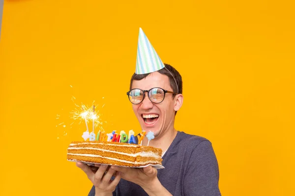 Loco alegre chico asiático joven con gafas sosteniendo una vela ardiente en sus manos y un pastel casero de felicitación sobre un fondo amarillo. Concepto de celebración de cumpleaños y aniversario . — Foto de Stock