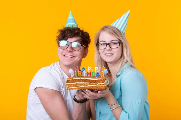 Wesoła Młoda para uroczy facet i cute girl w kapelusze papieru zrobić głupie twarz i trzymać w rękach ciasto z napisem urodziny stojący na żółtym tle. Pozdrowienia koncepcyjne i — Zdjęcie stockowe