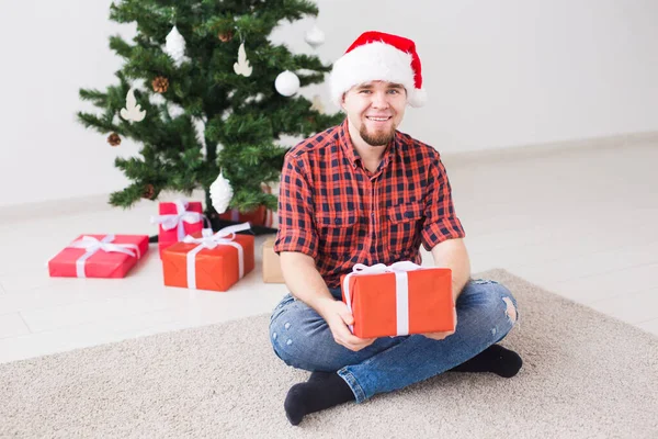 क्रिसमस और छुट्टियों की अवधारणा लिविंग रूम में घर पर उपहार पकड़ते हुए सांता टोपी में मजेदार आदमी — स्टॉक फ़ोटो, इमेज