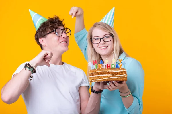 Jóvenes estudiantes alegres chica encantadora y buen chico en gorras de papel de saludo sosteniendo un pastel. Concepto de felicitación por el cumpleaños y aniversario —  Fotos de Stock