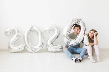 Bayramlar, şenlik ve parti konsepti - Beyaz arka planda, gümüş 2020 balon yakınında oturan mutlu çift. Yeni yıl kutlaması