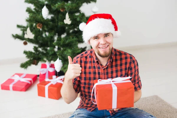 Weihnachts- und Feiertagskonzept - lustiger Mann mit Weihnachtsmütze, der zu Hause im Wohnzimmer ein Geschenk hält — Stockfoto