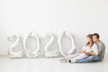 Bayramlar, şenlik ve parti konsepti - Beyaz arka planda, gümüş 2020 balon yakınında oturan mutlu çift. Yeni yıl kutlaması