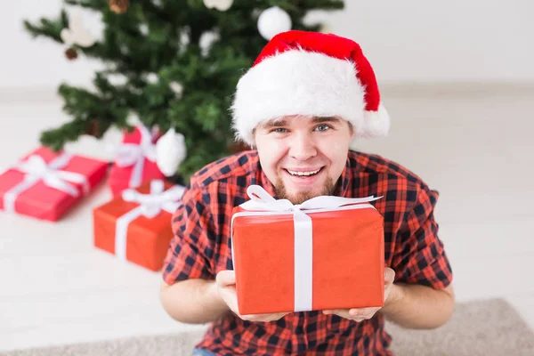 Kerstmis en feestdagen concept - Grappige man in santa hoed met een geschenk thuis in de woonkamer — Stockfoto