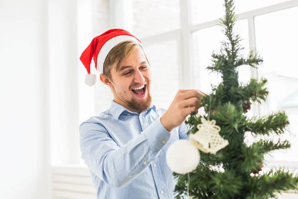 Homme heureux décorant arbre de Noël à la maison avec santa claus chapeau. Homme décorant l'arbre avec des boules pendant les vacances d'hiver . — Photo