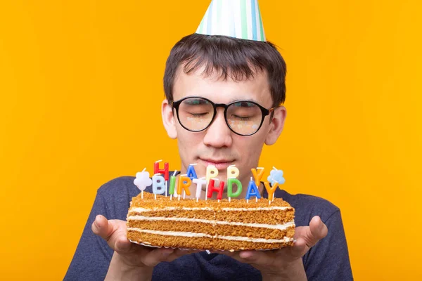 Портрет смешного позитивного парня в бумажном колпаке и очках с поздравляющим домашним тортом в руках на желтом фоне. Концепция и веселье и празднование . — стоковое фото