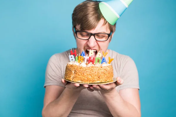 Loco joven divertido en gafas y sombreros de felicitación de papel celebración de pasteles feliz cumpleaños de pie sobre un fondo azul con copyspace —  Fotos de Stock