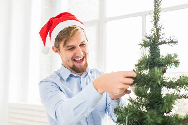 Un om fericit decorând pomul de Crăciun acasă cu pălărie de moș claus. Omul decorarea copac cu baubles în timpul vacanței de iarnă . — Fotografie, imagine de stoc