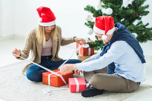 Boże Narodzenie i święta koncepcja - Młoda szczęśliwa para w kapeluszach Santa otwierających prezenty w domu. — Zdjęcie stockowe