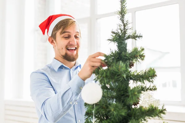 Gelukkig man versieren kerstboom thuis met kerstman hoed. Man versieren boom met kerstballen tijdens de wintervakantie. — Stockfoto